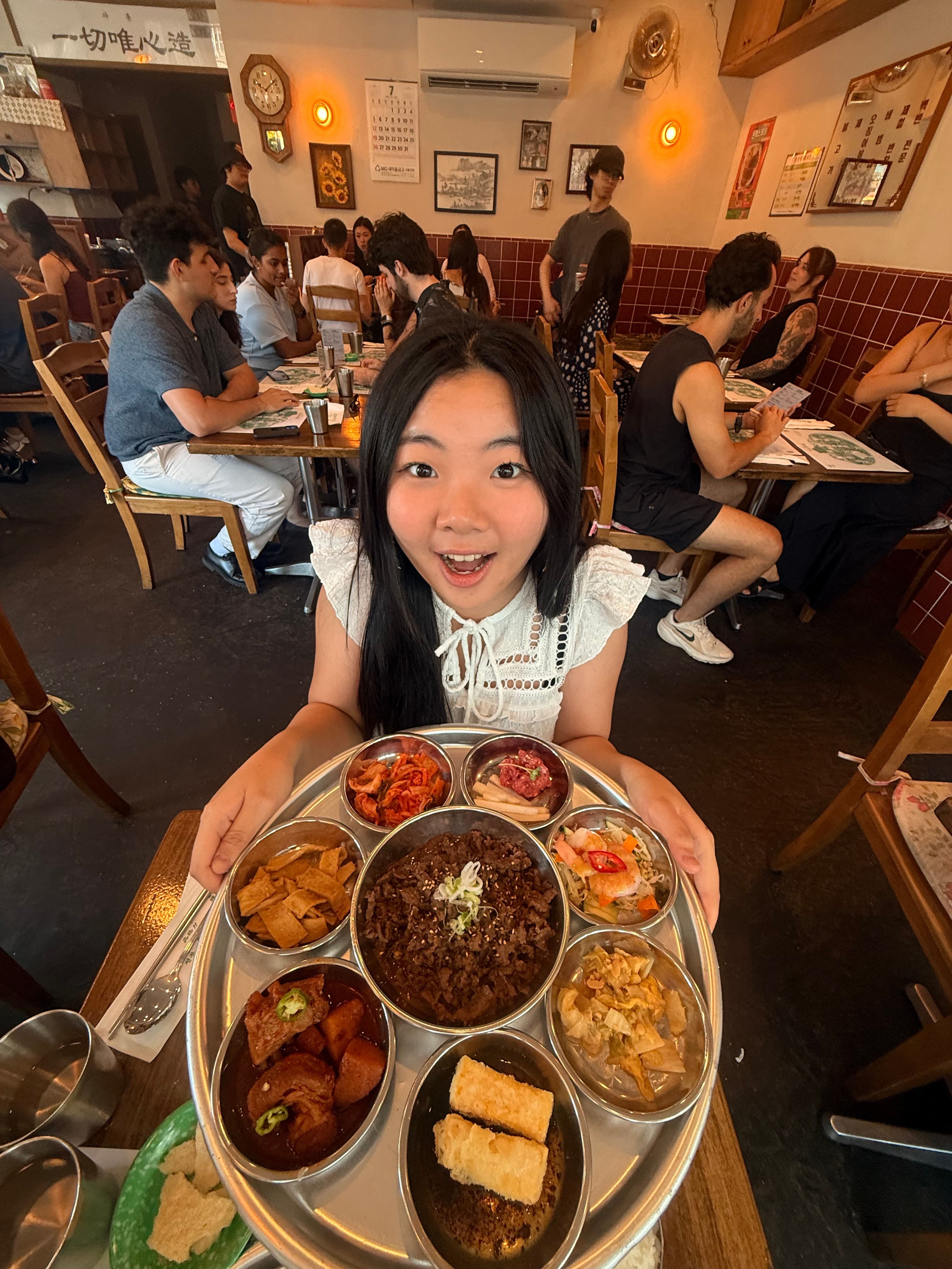 Jesslyn grinning wide-eyed as she holds up a round tray of Korean banchan around a bowl of bulgogi, in a busy restaurant