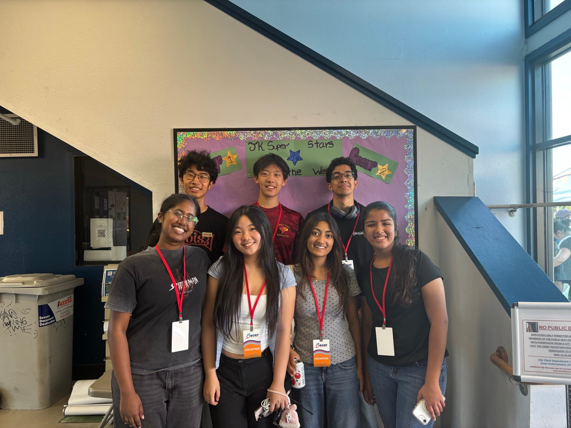 Jesslyn and seven Cal Blueprint teammates in BORP volunteer lanyards, posing in front of a bulletin board under a staircase