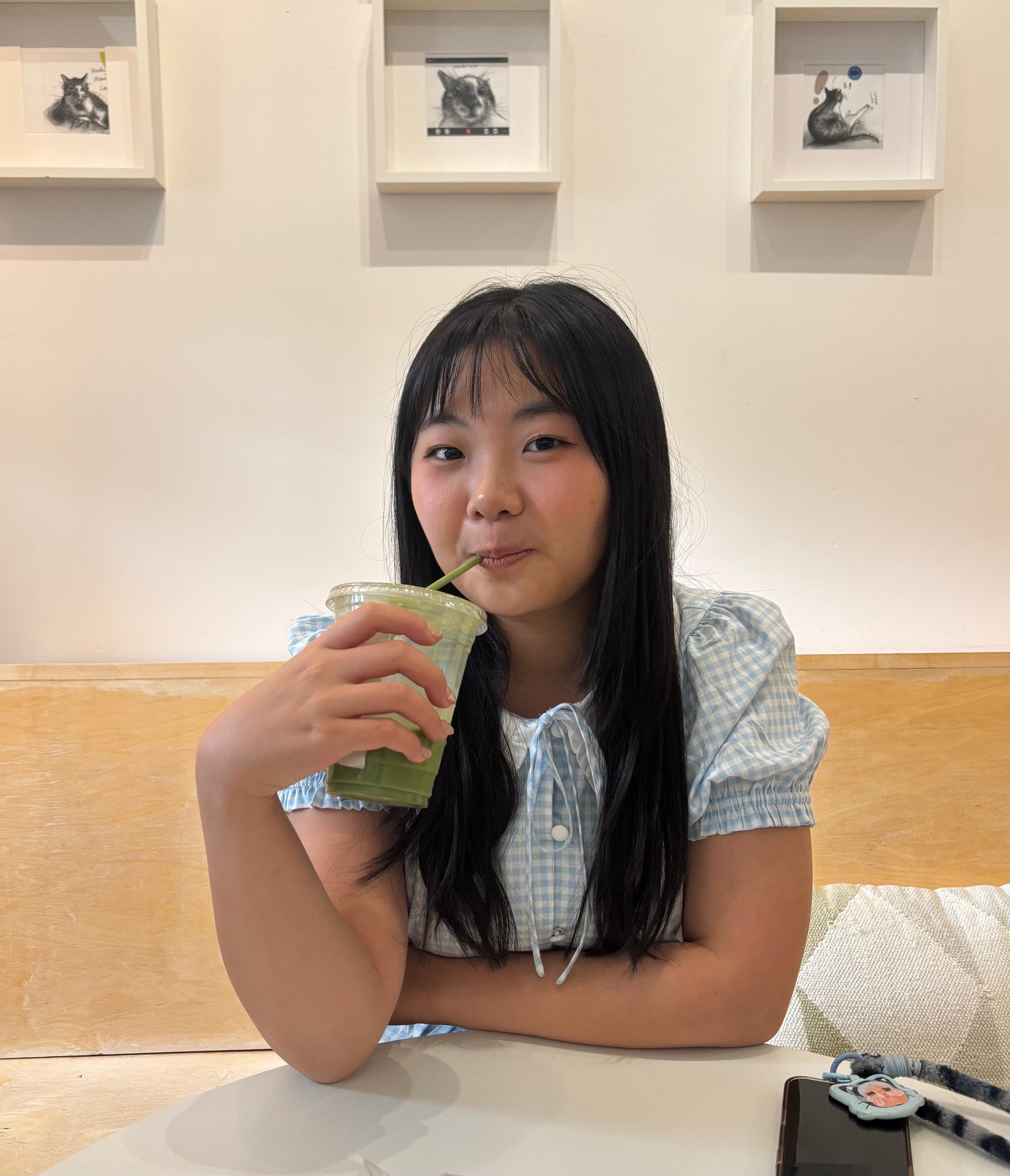 Jesslyn at a café table sipping an iced matcha, with framed cat drawings on the wall behind her