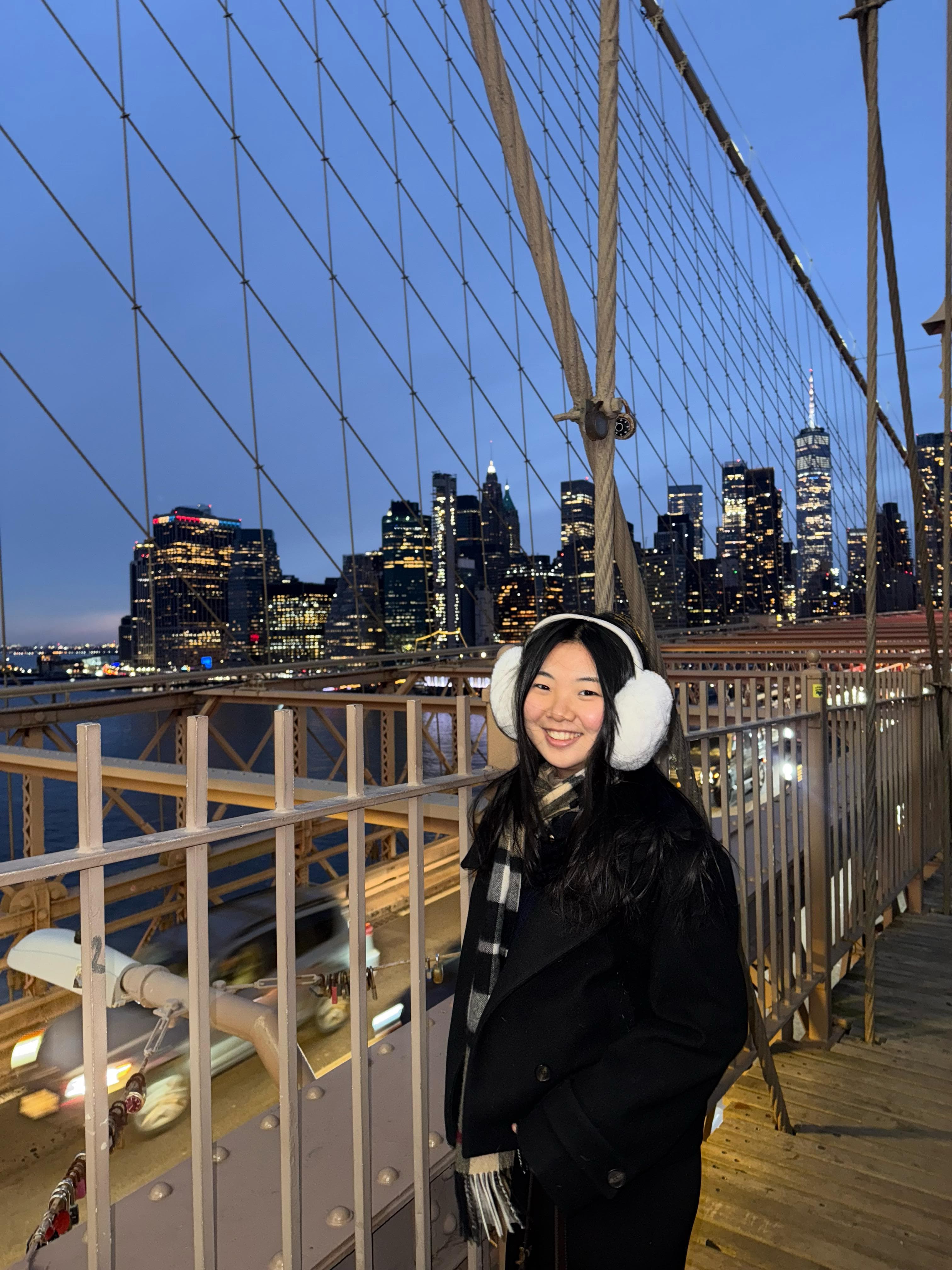 Jesslyn on the Brooklyn Bridge at night, wearing white earmuffs, with the Manhattan skyline behind her
